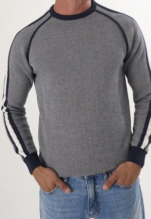 Mann trägt einen grauen Strickpullover mit marineblauen und weißen Streifen an den Ärmeln und blaue Jeans, die Hände in den Taschen.