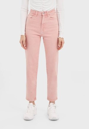 Woman wearing high-waist pink jeans, white ruched long-sleeve top, and white sneakers, standing against plain light background.