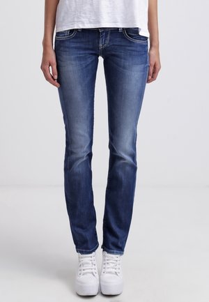 Jean skinny bleu foncé avec un devant décoloré, cinq poches et des détails de couture argentés, associés à des baskets blanches, se tenant sur un fond uni.