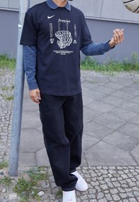 Camiseta negra de Nike con gráfico blanco de Brooklyn Nets, superpuesta a una camiseta de manga larga azul. Combinada con pantalones negros holgados y zapatillas blancas.