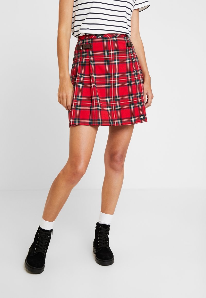 Roter karierter Rock mit Falten, braunen Schnallenakzenten, aus Stoff. Kombiniert mit einem gestreiften Oberteil und schwarzen Schnürstiefeln.