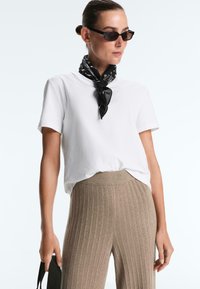 Kvinde iført hvid kortærmet skjorte, beige ribbede bukser, sort mønstret halstørklæde, slanke sorte solbriller, der holder en sort taske-rem.