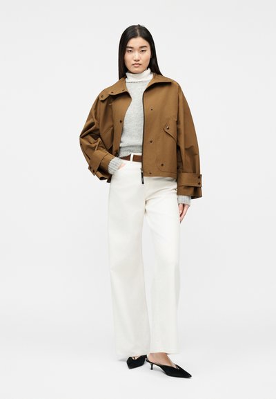 Γυναίκα με καφέ oversized μπουφάν, γκρι πουλόβερ, λευκό παντελόνι φαρδύ στα πόδια και μαύρες γόβες, στέκεται με το ένα χέρι στην τσέπη σε λευκό φόντο.