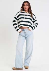 Ce sweat à capuche court présente des rayures vertes et blanches, une capuche avec cordon de serrage et des manches larges. Associé à un jean large bleu clair et des chaussures beiges.