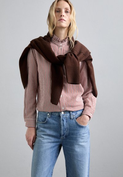 Κόκκινη και λευκή ριγέ πουκάμισο με κουμπιά, καφέ πουλόβερ ριγμένο στους ώμους, ανοιχτό μπλε τζιν παντελόνι με τσέπες. Λείες υφές, χαλαρή εφαρμογή.