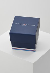 Navyblaues Uhrenetui mit glänzender Oberfläche, das den Text "TOMMY HILFIGER" und "Watches" in Weiß zeigt. Akzentuiert mit roten und blauen Streifen.