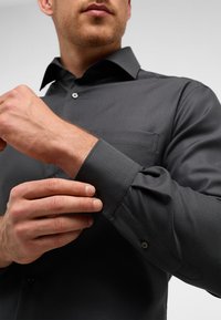 Graues Hemd mit Knöpfen aus strukturiertem Stoff, mit einem klassischen Kragen und einer Brusttasche. Die Manschetten sind aufgerollt und zeigen eine Knopfdetail.