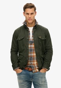 Chaqueta verde oliva con una camisa de cuadros con cuello debajo. Presenta dos bolsillos frontales, cierre de botones y una tela texturizada. Estilo casual.