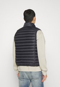 Mann in schwarzer, gesteppter Weste über einem beigen Sweatshirt und blauen Jeans, der mit dem Rücken zu einem weißen Hintergrund steht.