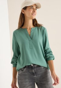 Türkise Bluse mit V-Ausschnitt und elastischen Bündchen, aus glattem Stoff. Kombiniert mit grauen Jeans aus Denim, die einen Knopfverschluss und Taschen haben.