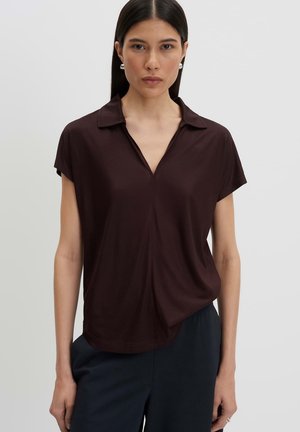 Vrouw met stijl donker haar draagt een donkerbruin kortemouw shirt met open kraag en donkere broek, staand tegen een effen lichte achtergrond.