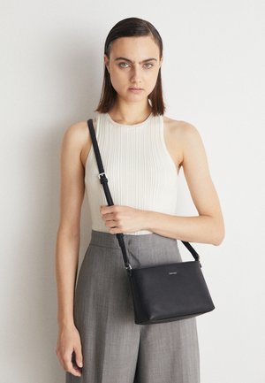 Jeune femme portant un haut blanc sans manches et un pantalon gris tient un sac bandoulière noir avec une lanière sur l'épaule.