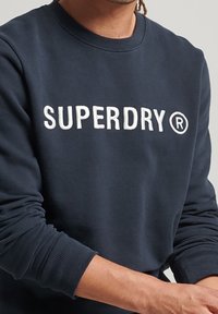Navy sweatshirt made of cotton blend, featuring ribbed cuffs and neckline. "SUPERDRY" logo in white across the chest.
