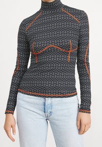 Långärmad topp i svart med vitt zigzag-mönster och orangea sömnadsdetaljer. Högkrage-design, åtsittande form, mjuk textur.