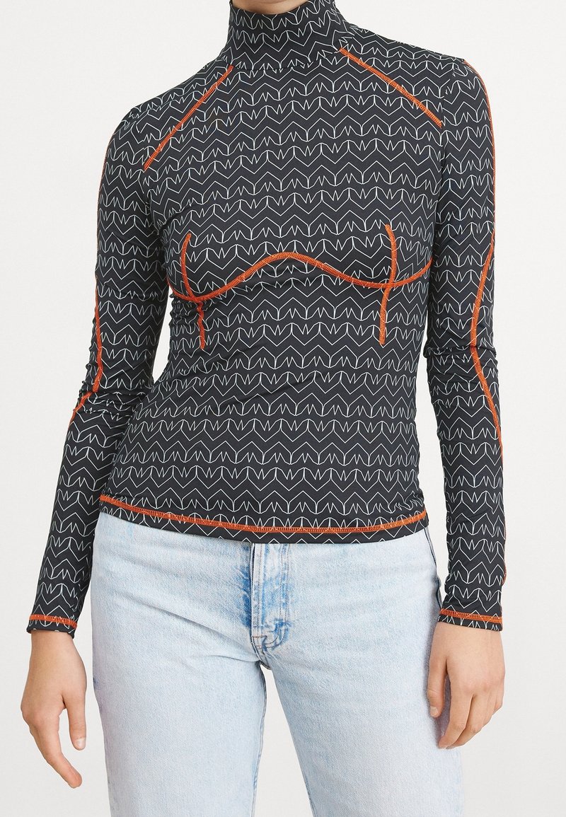 Långärmad topp i svart med vitt zigzag-mönster och orangea sömnadsdetaljer. Högkrage-design, åtsittande form, mjuk textur.