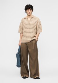 Νεαρός άνδρας που φοράει μπεζ πλεκτό πουλόβερ με κοντά μανίκια και φαρδιά καφέ παντελόνια, κρατώντας μια μπλε τσάντα tote, στέκεται μπροστά σε ένα απλό φόντο.