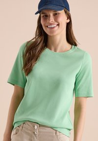 Femme souriante portant un haut côtelé vert clair à manches courtes, un pantalon beige à double boutons et une casquette de baseball bleu foncé.
