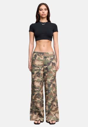 Joven mujer con camiseta negra corta, pantalones anchos de camuflaje y sandalias negras, de pie frente a un fondo claro y sencillo.