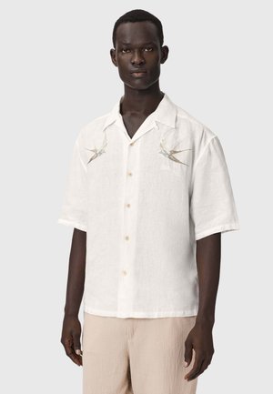 Homme portant une chemise blanche à manches courtes boutonnée avec des oiseaux brodés sur la poitrine et un pantalon beige clair, debout devant un fond uni.