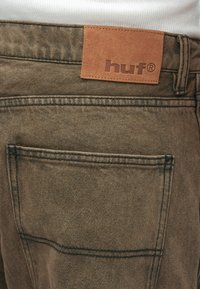 Καφέ τζην παντελόνι με δερμάτινη ταμπέλα με ανάγλυφη τη λέξη "huf." Διαθέτει πίσω τσέπη, ορατό ράψιμο και υφή επιφάνειας.