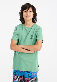Camiseta de algodón verde con mangas cortas que presenta un gráfico de una mano negra en el pecho. Combinada con pantalones cortos de patrones coloridos.