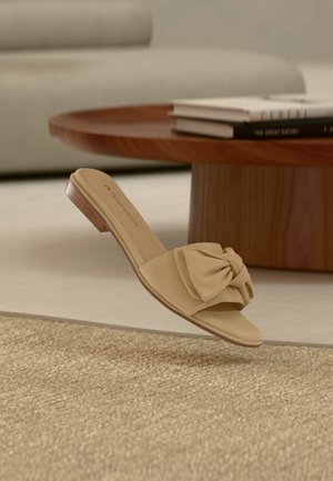 Sandale beige à enfiler avec un nœud décoratif noué sur le devant et un petit talon en bois, placée près d'une table basse ronde en bois avec des livres.