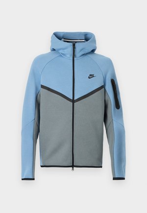 Casaco com capuz da Nike com uma combinação de cores azul e cinza, apresentando um fecho de correr frontal, um design em chevron e um bolso com fecho de correr na manga.