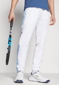 Bijele sportske hlače s plavim bočnim detaljima, elastičnim manžetama i logom. Model drži tenisku rekete te nosi bijele sportske tenisice.