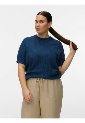 Hosszú, sötét hajú nő, aki fogja a lófarokját, kék texturált rövid ujjú pulóvert és bézs, húzózsinóros nadrágot visel, fehér háttér előtt áll.