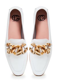 White leather slip-on shoes with a pointed toe. Features a chunky, tortoiseshell chain across the front, with a pink insole and subtle stitching.