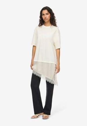 Jeune femme aux cheveux bouclés porte un t-shirt blanc oversize superposé à une jupe transparente avec ourlet en dentelle, un pantalon noir et des sandales blanches.