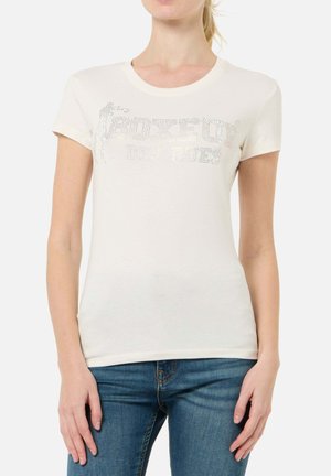 Donna che indossa una maglietta bianca aderente con scritta scintillante "BOXEUR DES RUES" e jeans blu, in piedi davanti a uno sfondo semplice.