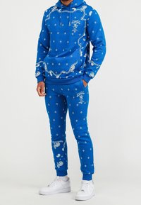 Niebieski zestaw: bluza z kapturem i spodnie dresowe z białymi wzorami paisley i kwiatowymi, kaptur ze sznurkiem, ściągacze na mankietach i w pasie, do tego białe trampki.