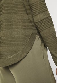 Olivgrüner Strickpullover mit geripptem Muster und abgerundetem Saum, kombiniert mit glatten olivgrünen Hosen mit Seitentaschen.