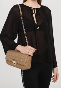 Beżowa pikowana torebka na ramię z złotym zapięciem i łańcuszkiem, w połączeniu z przezroczystą czarną bluzką z wiązanym dekoltem i szerokimi rękawami.