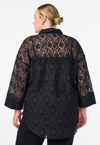 Čierna čipkovaná košeľa s 3/4 rukávmi, ktorá má priehľadný kvetinový vzor a pevnú čiernu časť na spodnej strane. Golier a okrúhly lem na konci.