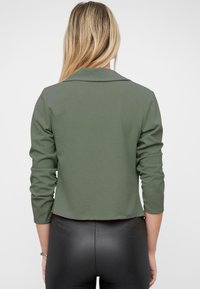 Chaqueta verde oliva recortada con un cuello entallado y mangas de tres cuartos, hecha de una tela suave y elástica.