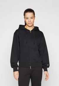 Crna zip-up dukserica s prednjim džepom, kapuljačom i vezicama. Ima mali logo na prsima i elastične manžete za udobno prianjanje.