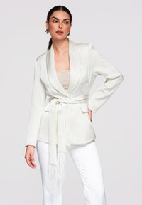 Femme aux cheveux foncés portant un blazer blanc ceinturé sur un haut beige et un pantalon blanc, posant avec une main sur la hanche devant un fond uni.