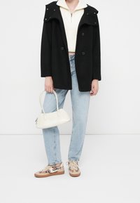 Person wearing a black coat, white zip-up sweater, light blue jeans, leopard-print sneakers, and holding a white handbag by one handle.