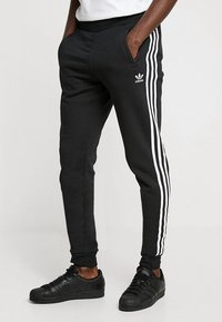 Svarta atletiska joggers med vit tre-stripesdesign längs sidan, elastisk midja och sidofickor. Bärs med svarta sneakers.