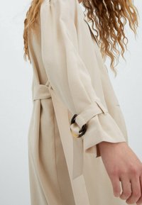 Un trench-coat beige en tissu lisse. Il présente une taille cintrée avec un accent en boucle et des manches retroussées. Design simple et droit.