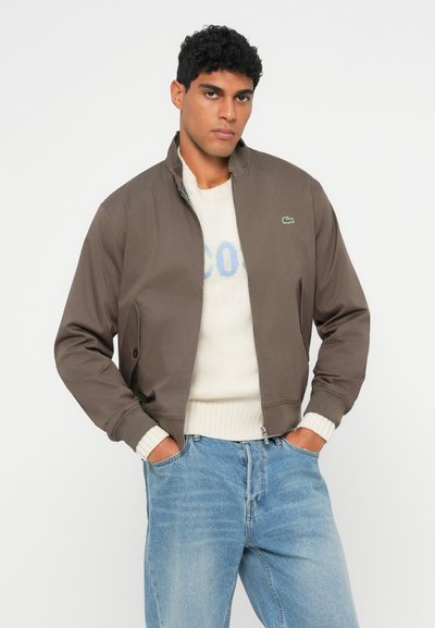 Homme aux cheveux noirs bouclés portant une veste zippée marron par-dessus un pull en maille blanc et un jean bleu clair, les mains dans les poches, se tenant devant un fond blanc.
