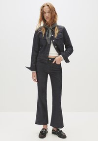 Mlada ženska v temnem denim jakni in razširjenih jeansih, beli kratki majici, sivoj šali, črnih loaferjih, stoji z eno roko v žepu.