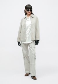 Hellgraue Wildlederjacke, weißes Satinhemd, Cargohosen, schwarze Handschuhe und übergroße schwarze Sonnenbrille, kombiniert mit braunen Stiefeln mit Absatz, stehend posiert.