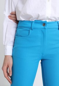 Pantalon en denim bleu avec une taille mi-haute, une fermeture à bouton sur le devant et des poches latérales. La texture est lisse avec une légèreélasticité.