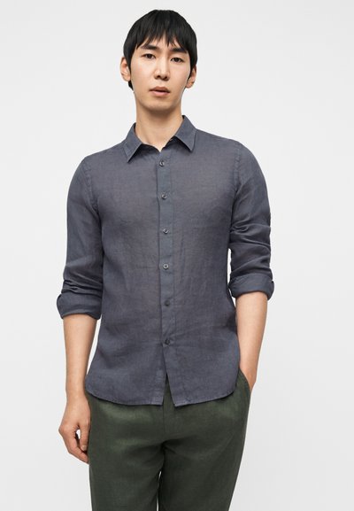 Σκούρο γκρι λινό πουκάμισο με κουμπιά στο μπροστινό μέρος, μακριά μανίκια διπλωμένα μέχρι τους αγκώνες και κανονικό γιακά. Φορεμένο με πράσινο παντελόνι.