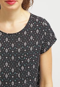 Mujer con una camisa de manga corta negra con patrones geométricos en blanco y verde azulado y un pequeño bolsillo en el pecho, sonriendo levemente.