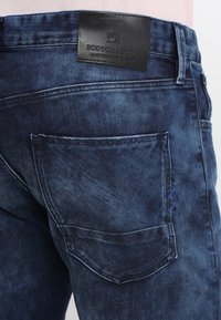 Jeans de mezclilla azul oscuro con un bolsillo trasero con costuras visibles y un parche de cuero negro con el logotipo de la marca en la cintura.