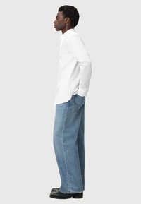 Camicia bianca a maniche lunghe con colletto a bottoni, abbinata a jeans in denim azzurro chiaro a vestibilità ampia e stivali neri, su uno sfondo neutro.